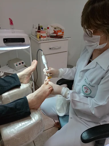 Felícia, podóloga do Espaço Felícia, atendendo uma cliente na sala de podologia — de jaleco, luvas e máscara, usando o micromotor.